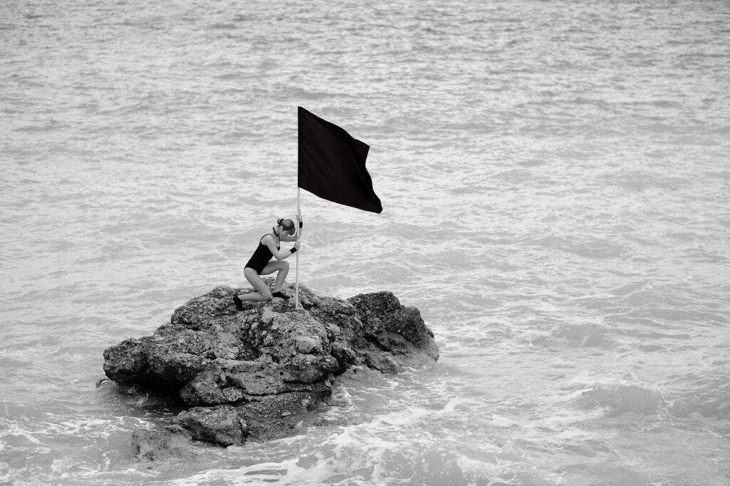 Auf dem Bild ist eine Frau im Schwimmdress, mitten im Meer auf einer kleinen schwimmenden Insel zu sehen, die eine Fahne in die Erde rammt.