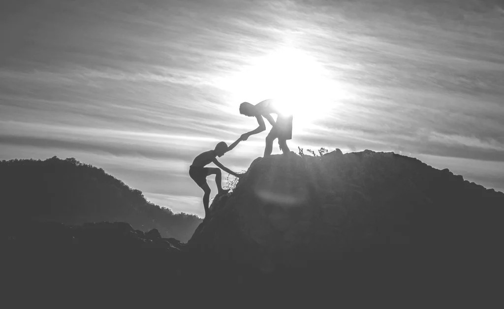 Auf dem Bild sind zwei junge Männer zu sehen, die sich gegenseitig unterstützen die Spitze eines Berges zu erreichen. Dabei hält ein Mann dem anderen die Hand hin und zieht diesen zu sich heran, um ihm ebenfalls das Erreichen der Bergspitze zu ermöglichen.
