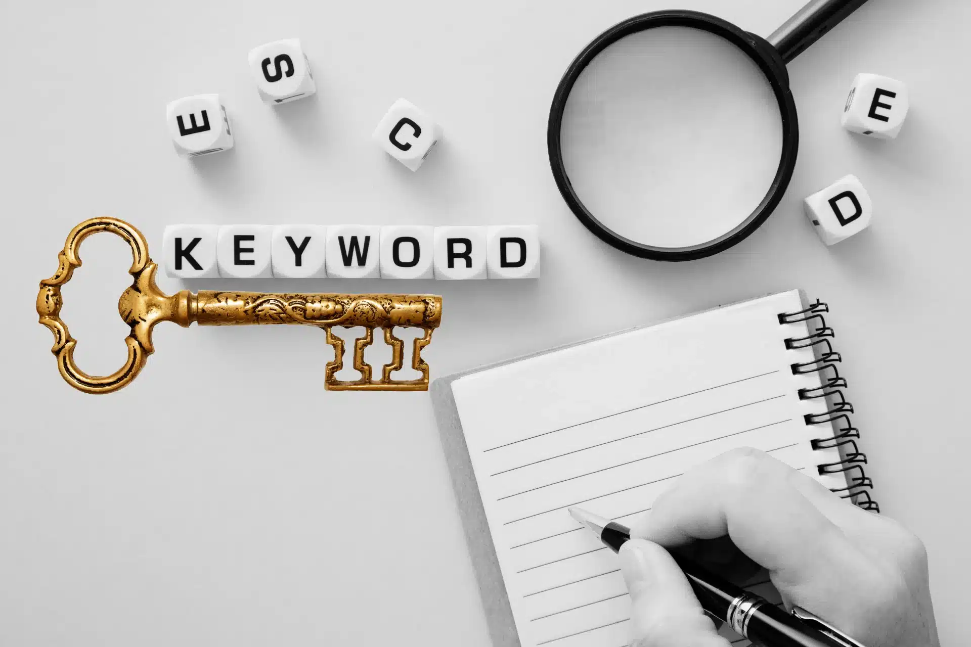 Das Bild zeigt die Hand einer Person, die eine Notiz auf einem Block vornimmt. Vor ihr auf dem Tisch liegen eine Lupe, ein goldener Schlüssel und Scrabble Buchstaben, die das Wort Keyword bilden.