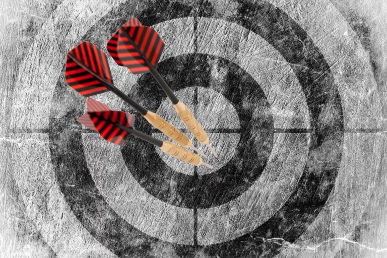 Das Bild zeigt eine Dart Zielscheibe, in deren Mitte drei rote Pfeile stecken. Es ist eine Allegorie für die Aussage: "Ziele erreichen"